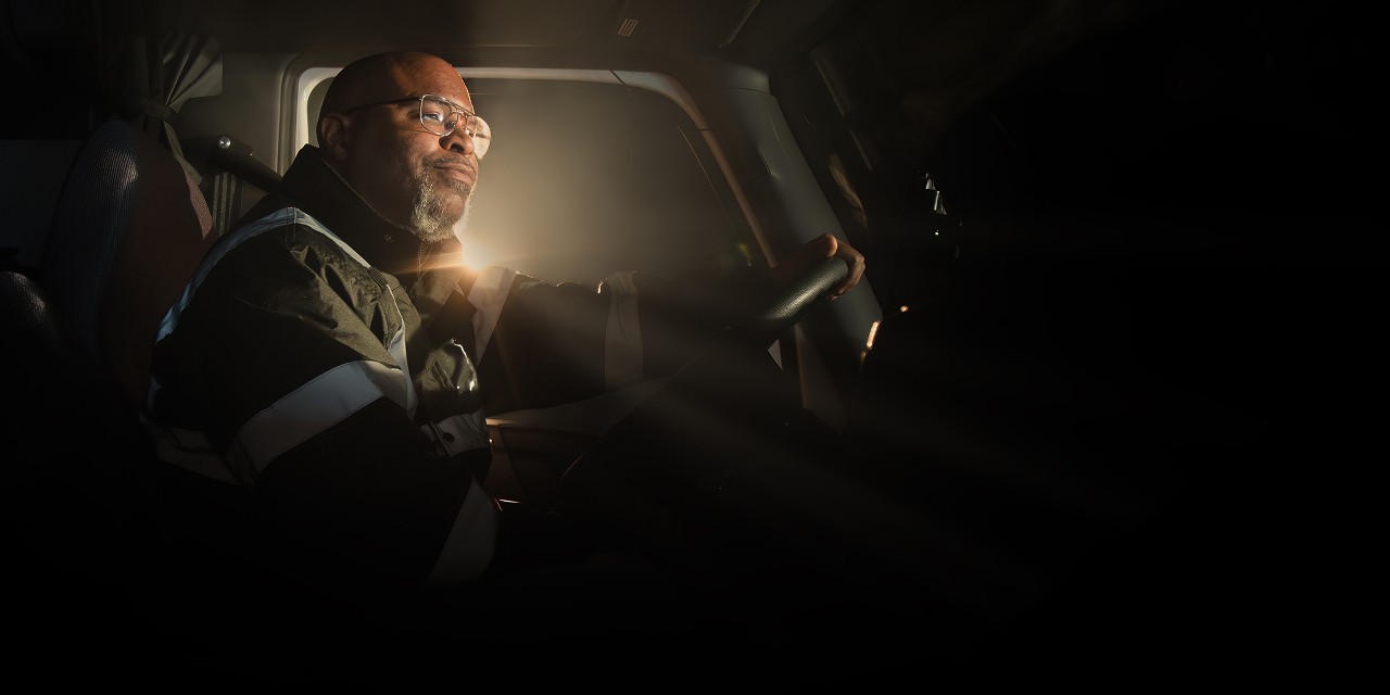 Truck driver seated inside the cab, holding the steering wheel with light shining through the windshield in a dark setting.
