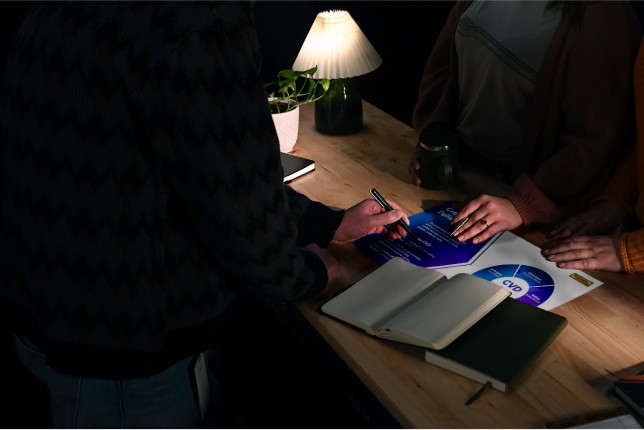 A team collaborating at a wooden desk in a dimly lit workspace, reviewing a printed marketing strategy document with charts. The table includes notebooks, a lamp, and a small plant.