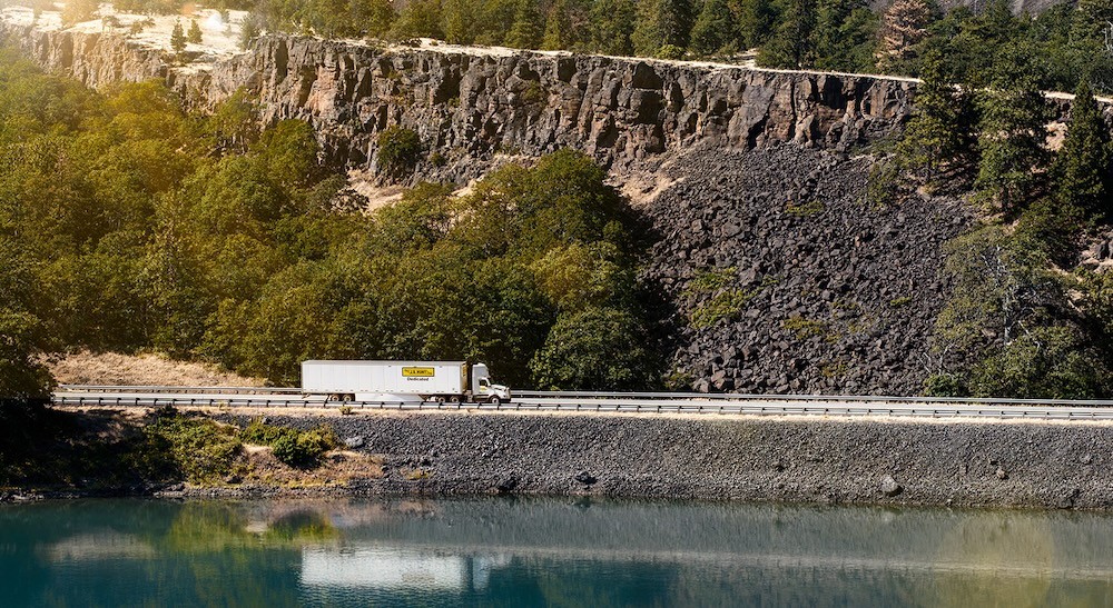 A white semi-truck with a yellow logo drives along a winding highway next to a calm body of water, with a lush green cliff face and dense trees in the background, illustrating efficient transportation and logistics in a beautiful landscape.