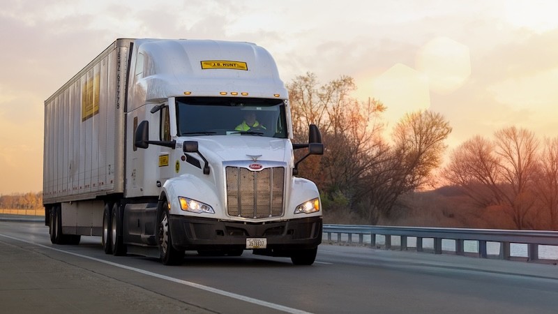 Front three-quarter view of a J.B. Hunt tractor trailer traveling on an open highway at sunset, featuring a white truck cab, box trailer, guardrails, and soft golden light with trees along the roadside.