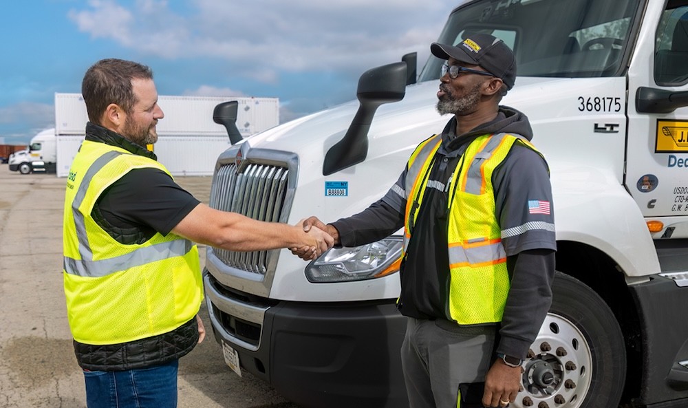 Two individuals wearing high-visibility safety vests shake hands in front of a parked commercial truck in an industrial yard, highlighting teamwork, safety, and professional collaboration.