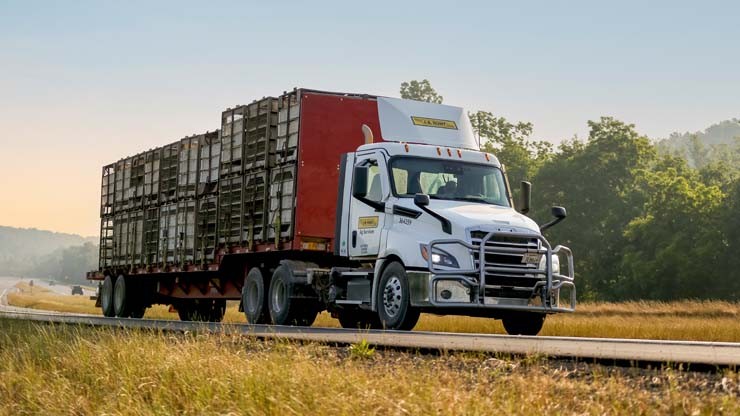 J.B. Hunt Dedicated Truck Transporting Agricultural Goods A J.B. Hunt truck hauling agricultural containers on a flatbed trailer, driving along a rural road with trees and open fields in the background under a clear sky.