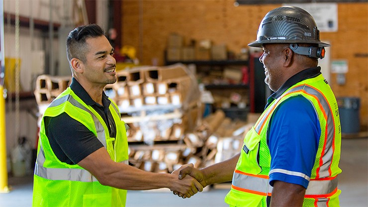 Two J.B. Hunt employees wearing safety vests and helmets shaking hands in a warehouse setting.