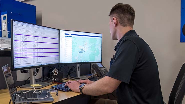 A logistics professional analyzing transportation data on dual monitors, featuring a spreadsheet and a digital map for route optimization.