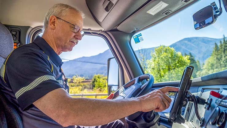 A truck driver using an onboard navigation and logistics system inside a J.B. Hunt truck, with scenic mountains in the background.