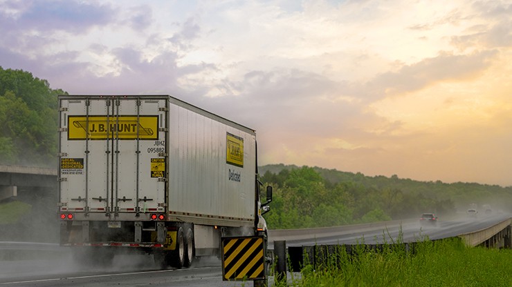 J.B. Hunt Dedicated Contract Services semi-truck driving on a misty highway surrounded by greenery during a colorful sunset