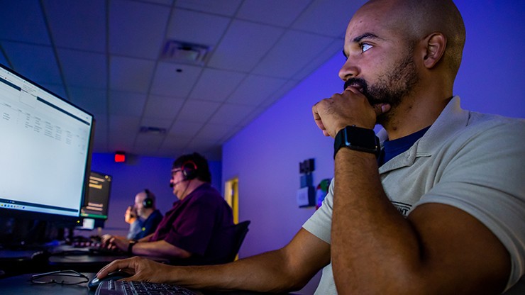 A cybersecurity analyst in a dimly lit control room closely monitors computer screens displaying data.