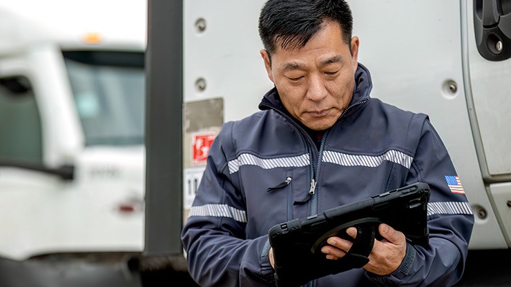 A truck driver in a uniform using a rugged tablet for logistics and fleet management, with trucks in the background.
