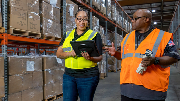managed_logistics_warehouse_employee Two warehouse employees in high-visibility safety vests discussing logistics while working in a warehouse environment. One is holding a tablet and gesturing, while the other listens, holding a scanner and gloves. They are surrounded by pallets of packaged goods on metal shelving.