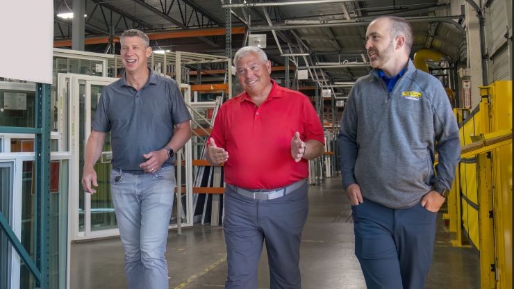j.b. hunt employees meet with milgard windows representative in warehouse Three men walk through a window manufacturing facility. The man in the center wears a red polo shirt and gestures while smiling, flanked by two men—one in a gray polo shirt and the other in a gray quarter-zip pullover with the J.B. Hunt logo.