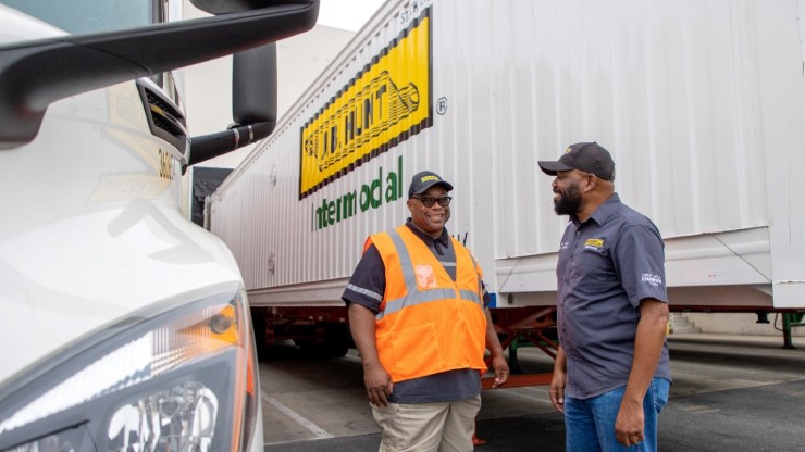 Two men in J.B. Hunt uniforms stand and talk in front of an intermodal trailer, one wearing an orange safety vest and the other in a gray company shirt, with a truck visible in the foreground.