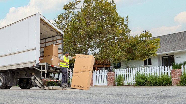 Delivery worker unloading large boxes from a J.B. Hunt Final Mile truck using a liftgate in front of a residential home