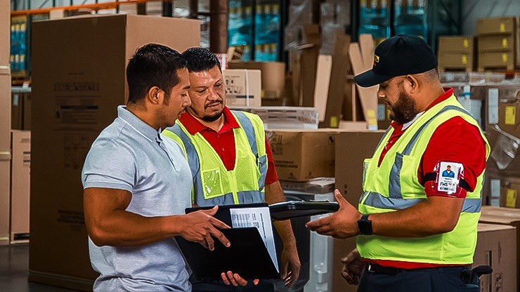 Three warehouse workers in safety vests discussing logistics and reviewing shipping documents inside a storage facility filled with boxes.
