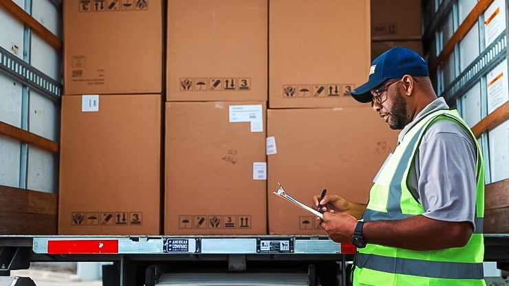A warehouse worker in a high-visibility vest and cap checking inventory on a clipboard next to an open truck loaded with large boxes.