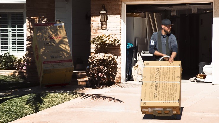 A delivery worker using a hand truck to move a large appliance box toward a residential home with an open garage.