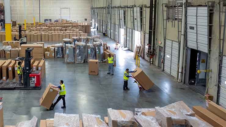 Warehouse with workers in safety vests moving large boxes near loading bays.