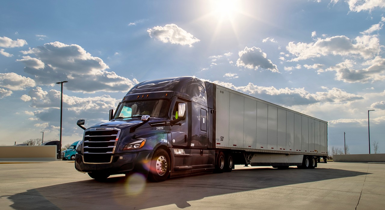 A dark-colored semi-truck with a white trailer parked on a concrete lot under a bright blue sky with scattered clouds, representing reliable truckload shipping and freight brokerage solutions.