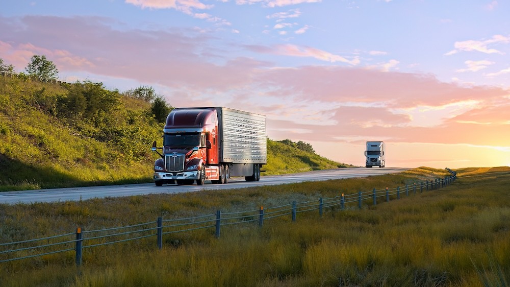 Two semi trucks traveling on a rural highway during sunset with open fields and rolling hills, representing long-haul logistics and freight transportation