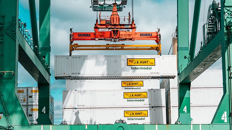 Multiple J.B. Hunt intermodal containers are stacked at a shipping port, with an orange crane lifting one of the containers. The scene is framed by green port equipment and a bright sky.