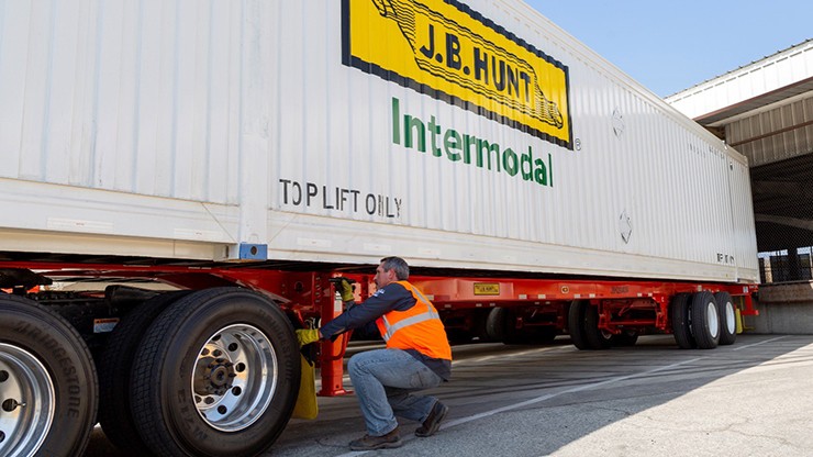 A worker wearing an orange safety vest and jeans secures a J.B. Hunt intermodal container to a chassis in a loading area. The white container features the J.B. Hunt logo.