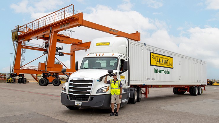 A J.B. Hunt intermodal truck parked at a container yard, with a driver in a safety vest standing beside it. Orange cargo cranes and shipping containers are visible in the background under a partly cloudy sky.