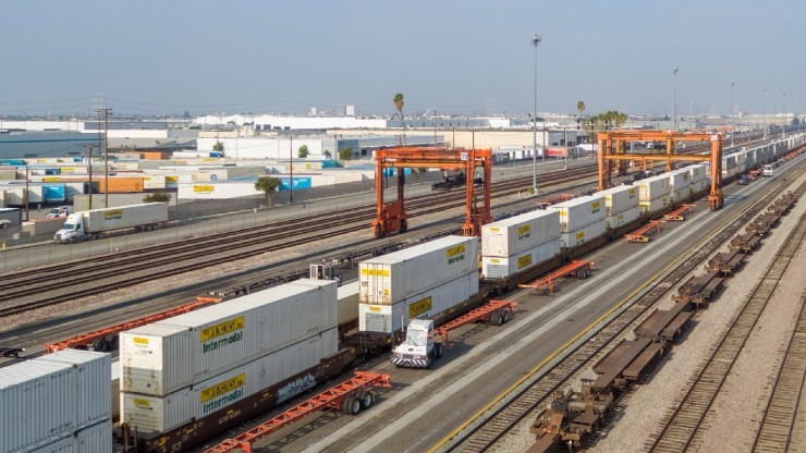 A busy intermodal rail yard featuring a long container train loaded with yellow and white trailers, with gantry cranes and industrial buildings in the background.