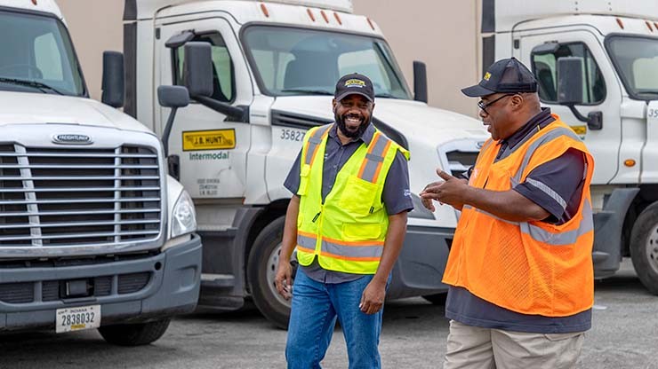 Two truck drivers wearing safety vests chatting and laughing in front of parked semi-trucks.