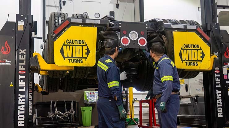 Two truck technicians inspecting the undercarriage of a lifted semi-truck in a maintenance shop.