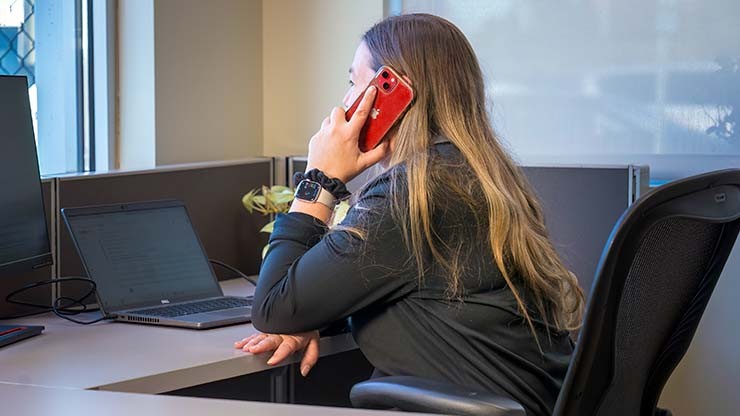 A logistics office worker speaking on a red phone while working on a laptop at her desk.