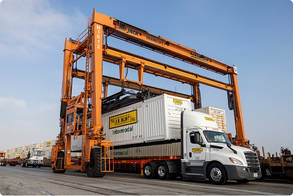 A white J.B. Hunt semi-truck hauls an intermodal container, which is being lifted by a large orange gantry crane at a busy logistics facility.