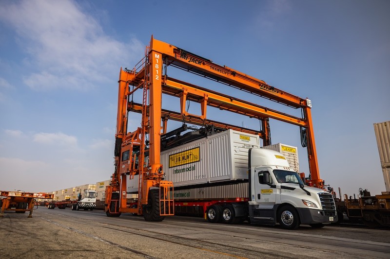 Large orange overhead crane lifting an intermodal container from a white semi‑truck inside an active rail yard, with trailers and equipment lined up in the background under a clear sky.