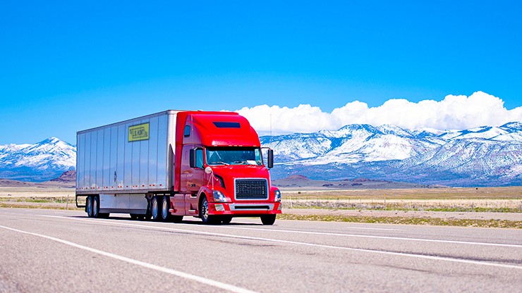 Large beautiful modern classic-modern red truck with a high cab and trailer on a flat stretch of highway on a background of snow-capped mountain ranges, drowning in the clouds and clear blue sky