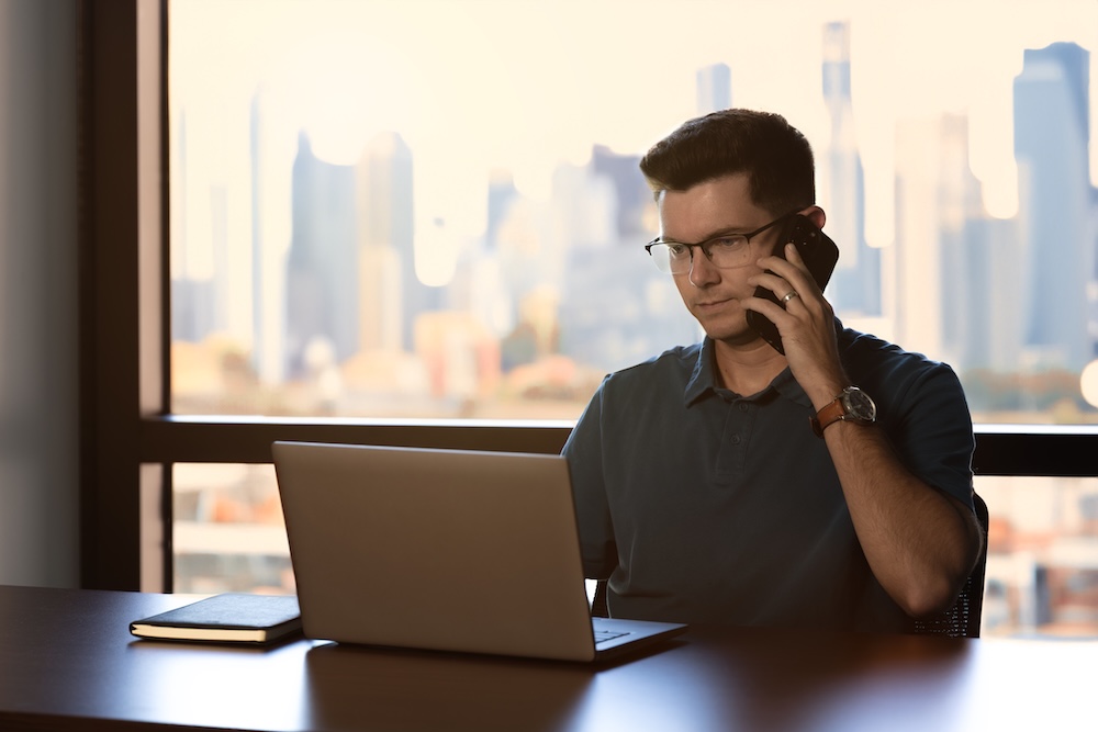 Individual seated at an office desk with a laptop and notebook while speaking on the phone, city skyline visible through large windows, representing logistics coordination.
