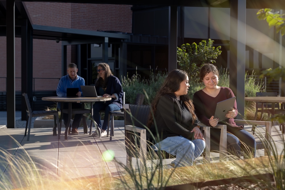 Two pairs of individuals sitting outdoors at tables in a landscaped patio area, engaged in conversation and working on laptops, representing a casual and collaborative workplace.