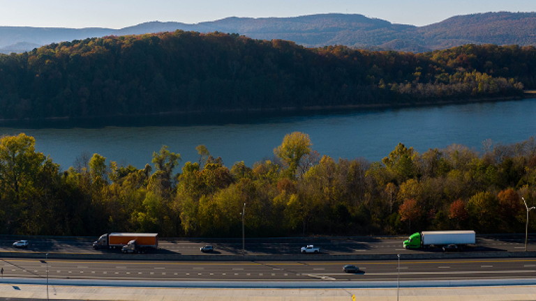 Semi-trucks and cars traveling on a highway beside a river, with forested hills and mountains in the background.