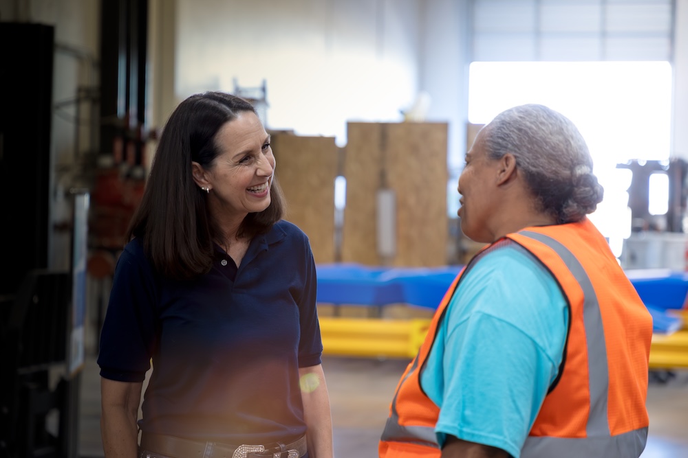 Two people conversing indoors near industrial equipment, one in safety gear and the other in supervisory attire, highlighting customer-focused operations.