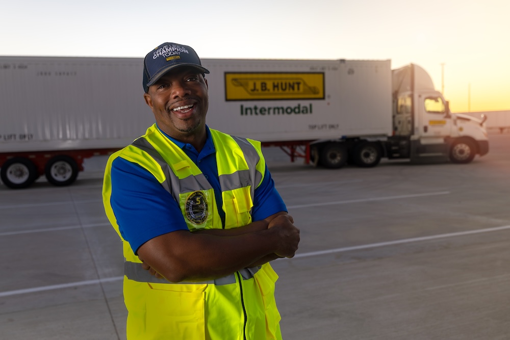 Person wearing high-visibility safety gear standing near intermodal containers labeled J.B. Hunt, representing skilled drivers in freight handling.