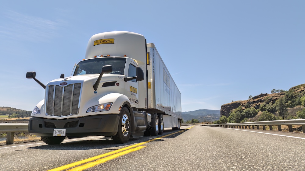 J.B. Hunt tractor‑trailer driving on an open highway under a clear sky, representing nationwide freight transportation and logistics services.