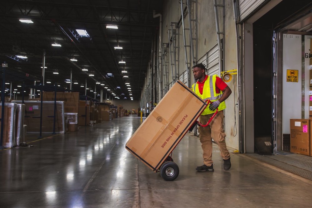 J.B. Hunt employee in safety vest using hand truck to move box in warehouse with loading docks, showing logistics operations.