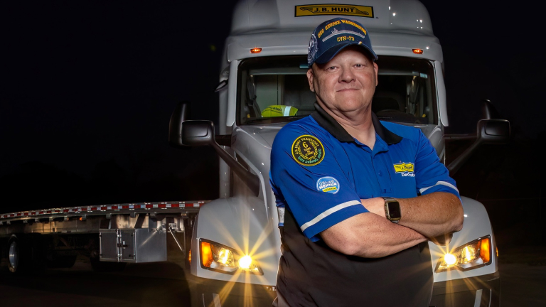 Truck driver in uniform standing with arms crossed in front of a J.B. Hunt semi-truck at night.