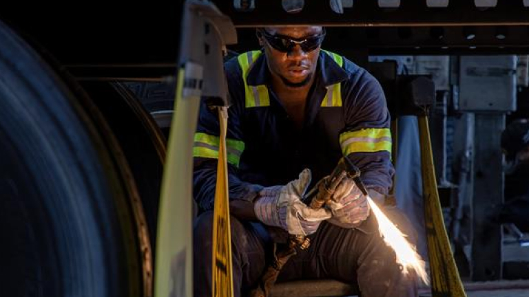 Technician wearing safety gloves and reflective stripes using a torch to perform maintenance under a trailer.