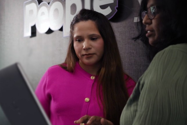 Two women discussing work in front of a laptop, with a “People” sign visible in the background. One woman is wearing a pink blazer, and the other is dressed in green.