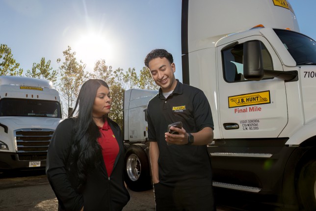 Two J.B. Hunt employees stand in front of Final Mile delivery trucks, reviewing information on a smartphone under a bright, sunny sky.