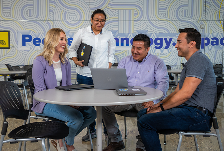 Four people sit around a small round table smiling at a laptop