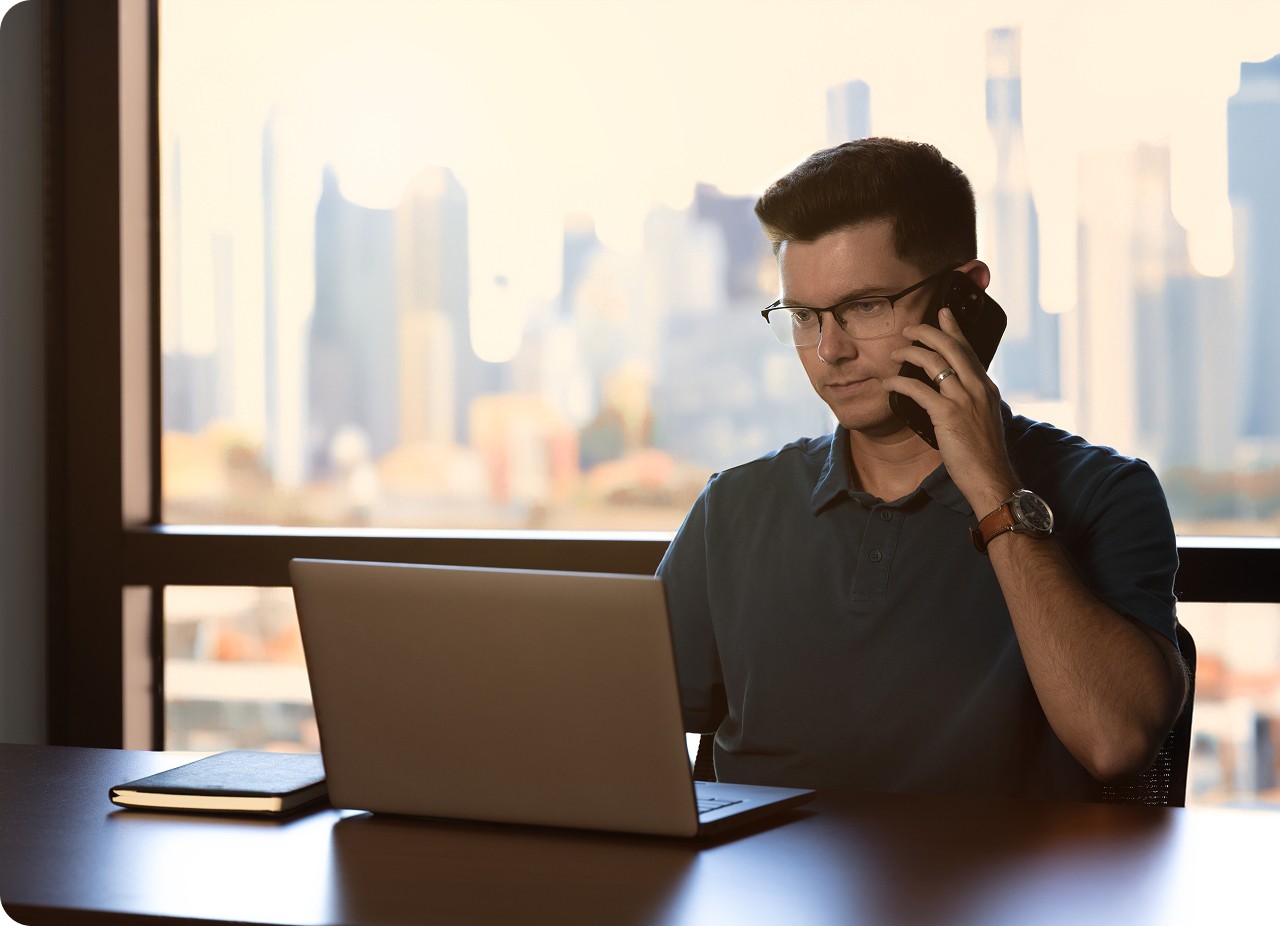 Freight agent sitting at a desk with a laptop and notebook, talking on the phone in front of a large window overlooking a city skyline.