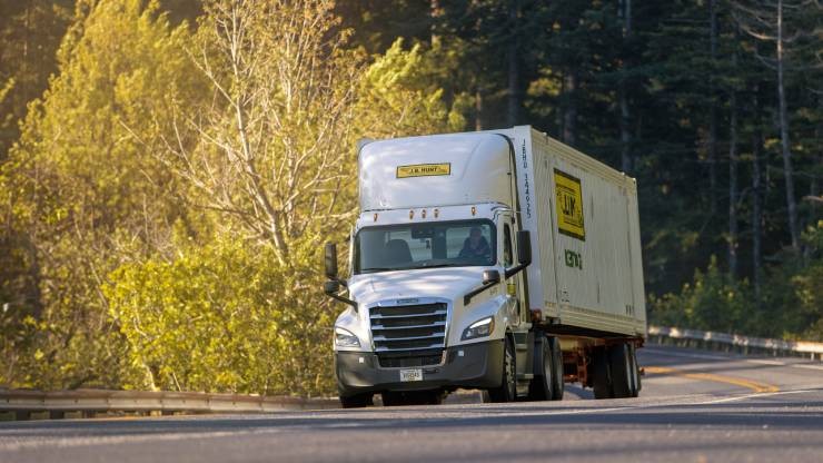J.B. Hunt Semi-Truck Driving on a Curved Road in a Forested Area A J.B. Hunt semi-truck hauling a shipping container navigates a curved road surrounded by trees and greenery.