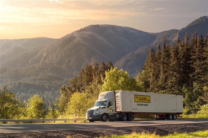 A J.B. Hunt intermodal truck travels along a highway surrounded by tall evergreen trees and distant mountains under warm, early morning light.