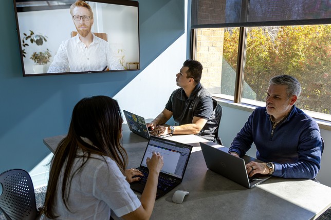 Jimi meeting with two people at a conference table and one attendee on a screen. 
