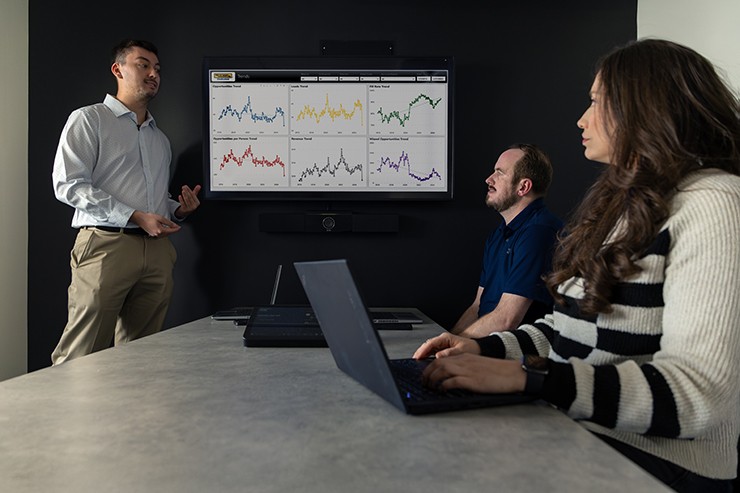 A business professional presents data charts on a large screen to two colleagues in a modern conference room.