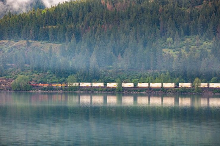 Intermodal freight train with stacked shipping containers traveling along a lakeside rail line, with forested hills and a clear water reflection in a misty, mountainous landscape.